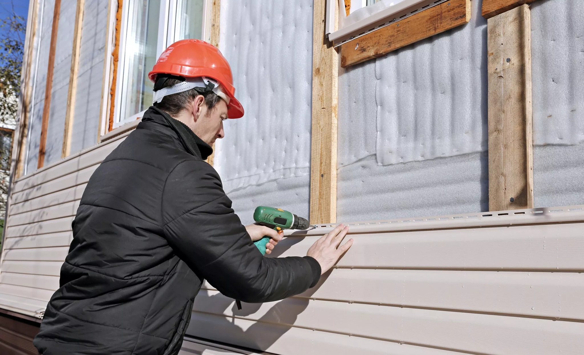Bozeman Vinyl Siding Installers Near Me securing the latest row of vinyl siding. A construction worker in an orange hard hat and black jacket uses a cordless drill to install tan vinyl siding on a house. Professional installation services by Wegner Roofing & Solar.