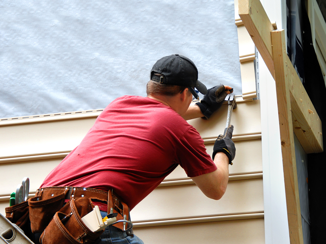 Bozeman Vinyl Siding Installers Near Me working on a residential siding project. A contractor wearing a red shirt, black hat, and work gloves is using a siding tool to fasten a tan vinyl panel to the house frame and house wrap. Siding installation by Wegner Roofing & Solar.