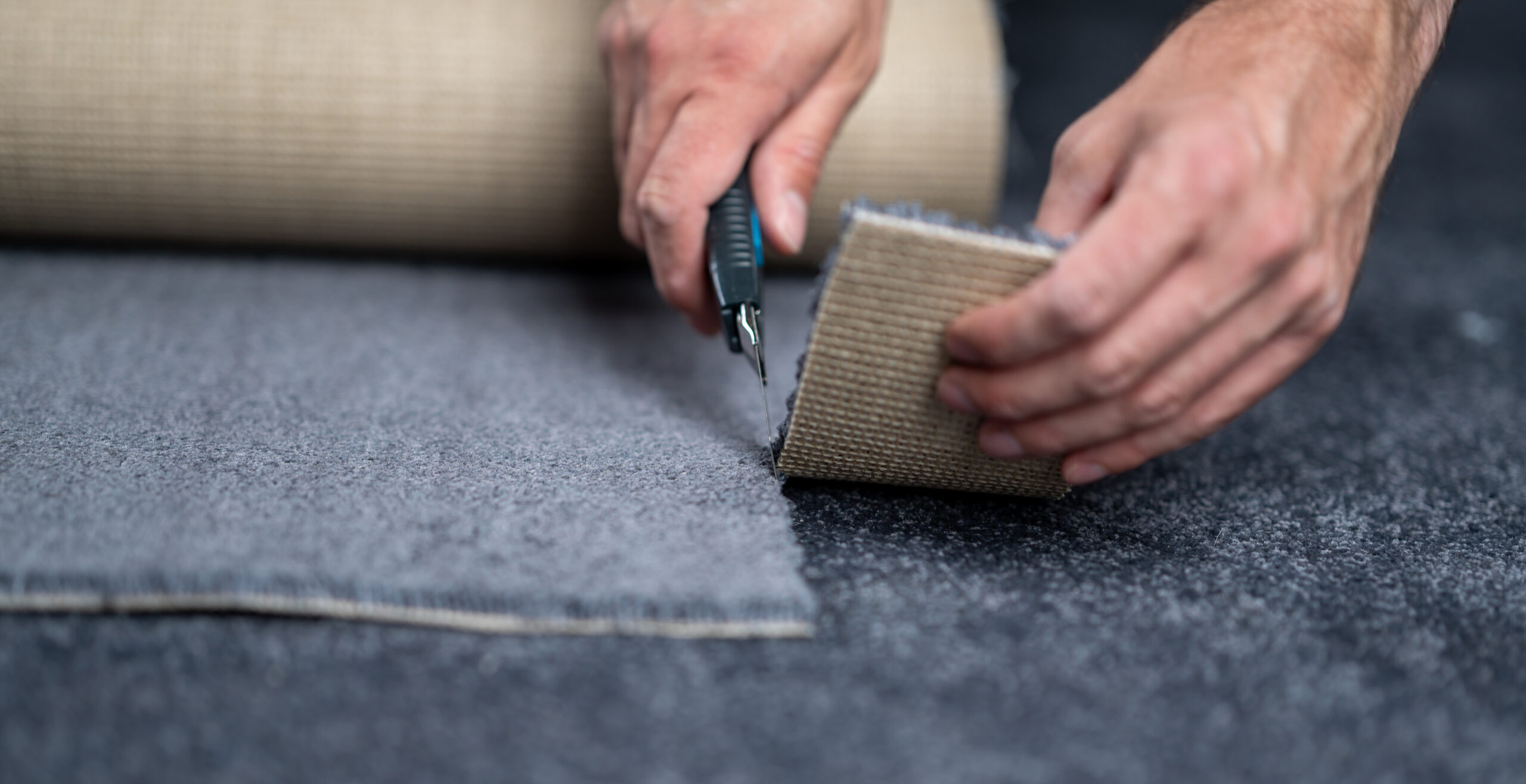 Professional carpet installer trimming a seam using a utility knife, showing a transition between dark gray and tan rolled carpet during Carpet Installation Roswell GA by Peachy Flooring Solutions.