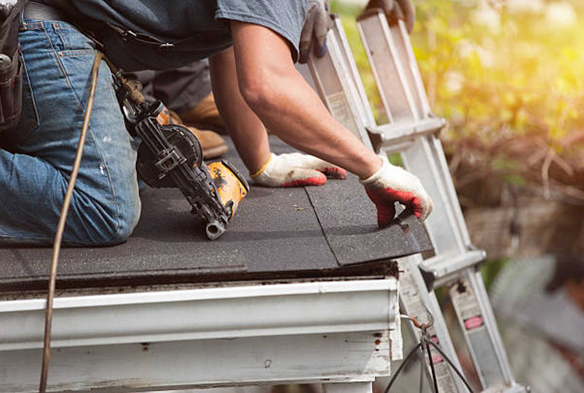 A close-up shot of a contractor wearing gloves and blue jeans, kneeling on a roof while operating a nail gun near the white gutter. The worker is installing or repairing asphalt shingles near the edge of the roof next to a ladder. This illustrates roof work related to Carver MN Gutter Damage by Pro24 Contracting, Inc.