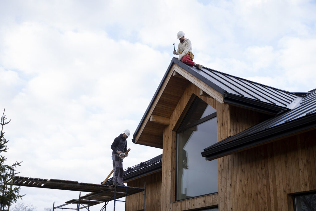 Two contractors working on a newly installed black metal roof on a modern wood-sided house, showing the progress of a Carver MN Roof Replacement by Pro24 Contracting, Inc. One contractor is on scaffolding, and the other is sitting on the peak of the roof.