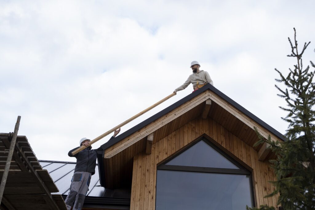 Two contractors wearing hard hats installing a wooden beam on the peak of a modern wood-sided house during a Carver MN Roof Replacement project by Pro24 Contracting, Inc.