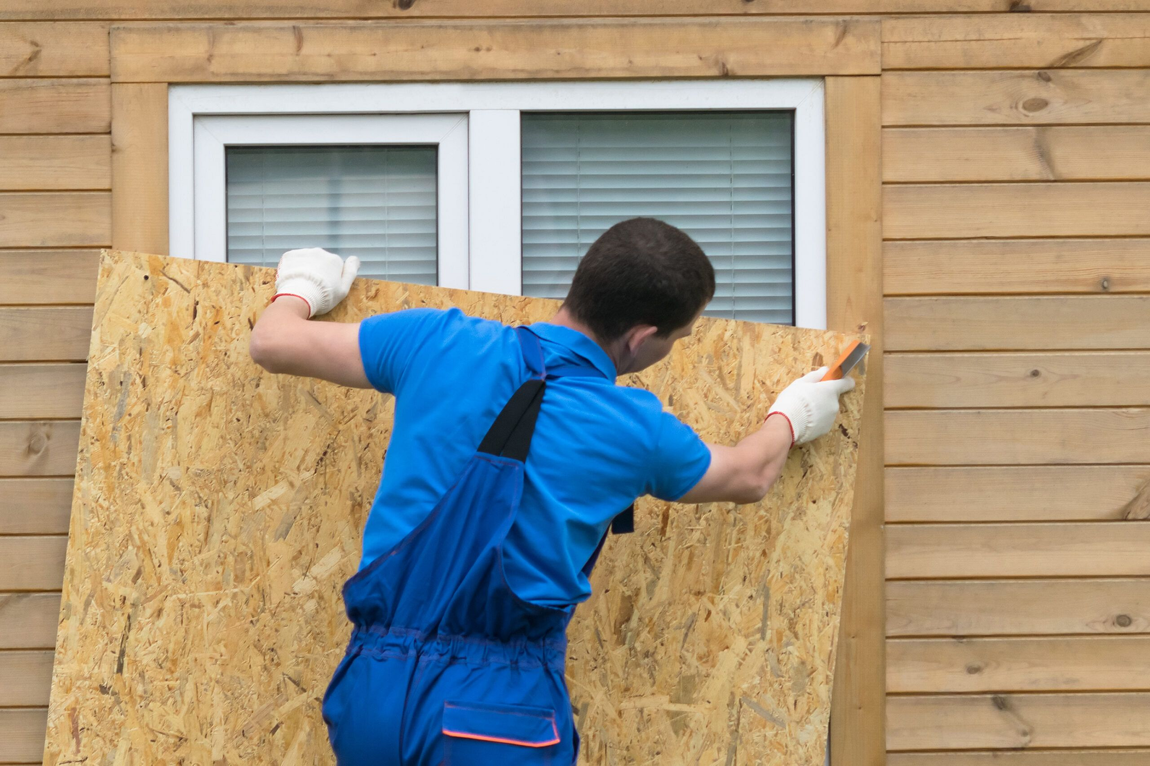 Carver MN Siding Contractor by Pro24 Contracting, Inc. worker in blue overalls securing a sheet of OSB or plywood over a window on a wooden-sided residential house, likely for temporary storm protection or repair prep.