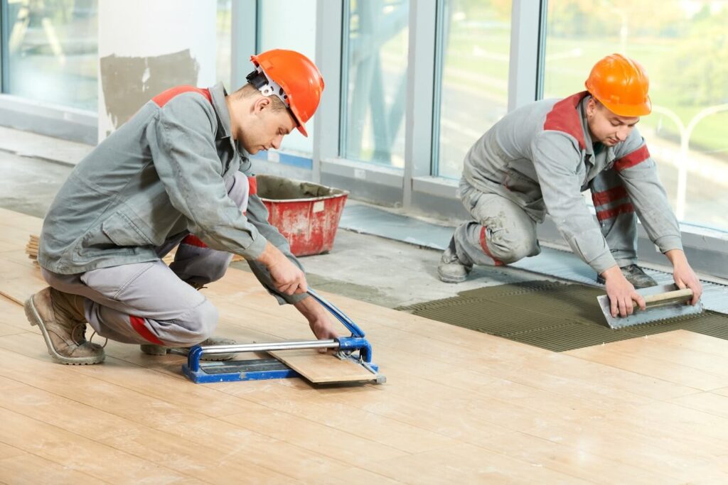 Two flooring contractors perform a Commercial Flooring Installation Roswell GA, one cutting a wood-look plank tile while the other spreads adhesive on the subfloor. Quality work by Peachy Flooring Solutions.