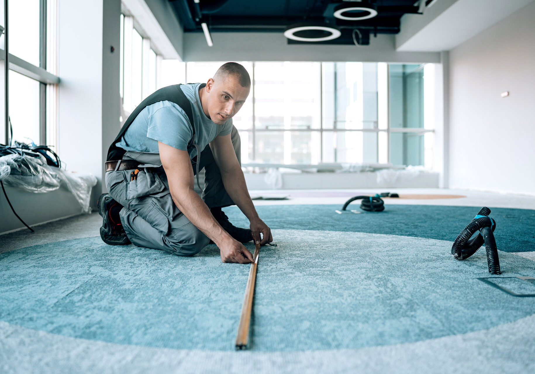 Commercial Flooring Installation Roswell GA - A worker measures a section of light blue commercial carpet being installed in a modern, sunlit office space, ensuring a precise fit. Project by Peachy Flooring Solutions.