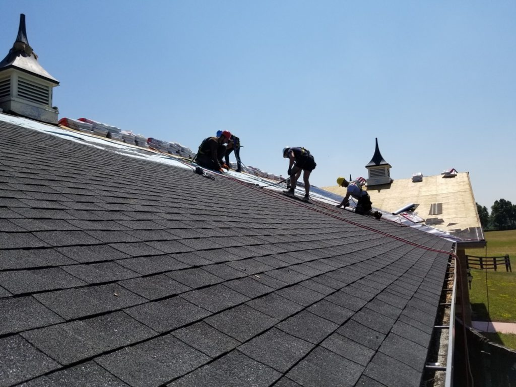 Roofing crew installing black asphalt shingles on a steep-sloped residential roof with decorative cupolas under a clear blue sky. The workers are wearing safety harnesses, and piles of new roofing materials are visible near the ridge. Forrest City NC Residential Roofing Services by Burish Builder Asheville.