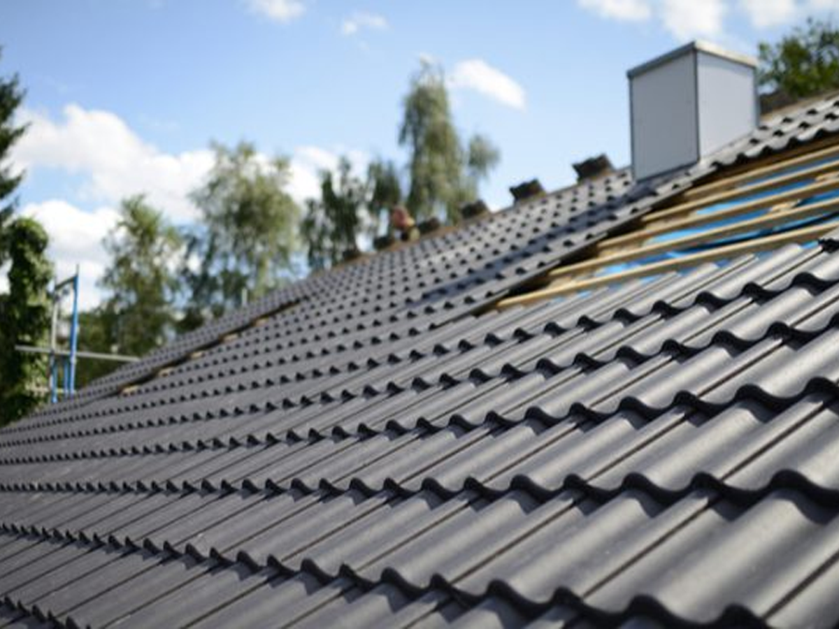 Close-up view of a partially completed roof installation featuring dark gray metal tiles with a pronounced wave pattern. The new roofing material is covering a section where wooden battens are visible against a backdrop of trees and a blue sky. Forrest City NC Residential Roofing Services by Burish Builder Asheville.