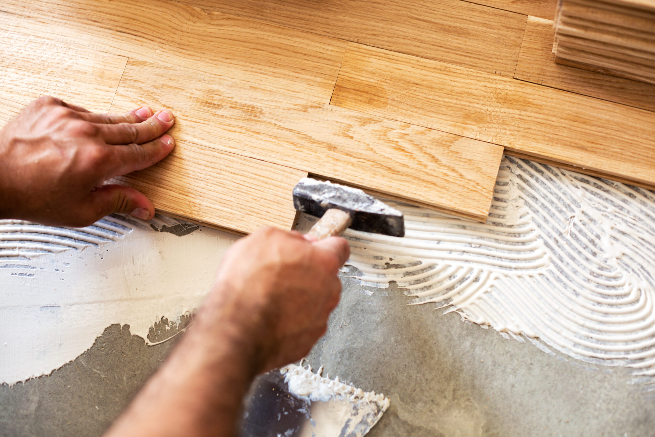 Close-up view of a professional installer using a hammer to tap a plank of hardwood flooring into place over adhesive, showcasing the detailed work of Hardwood Floor Installation Marietta GA by Peachy Flooring Solutions.