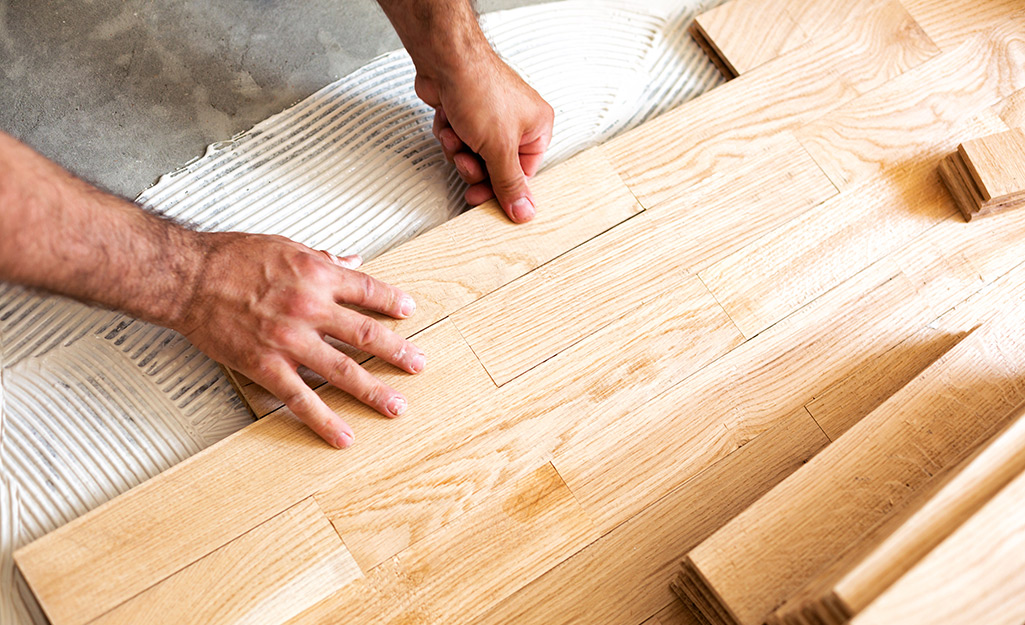 A contractor's hands placing a plank of light-colored, unfinished hardwood flooring onto an adhesive-covered subfloor during a Hardwood Floor Installation Marietta GA project by Peachy Flooring Solutions.