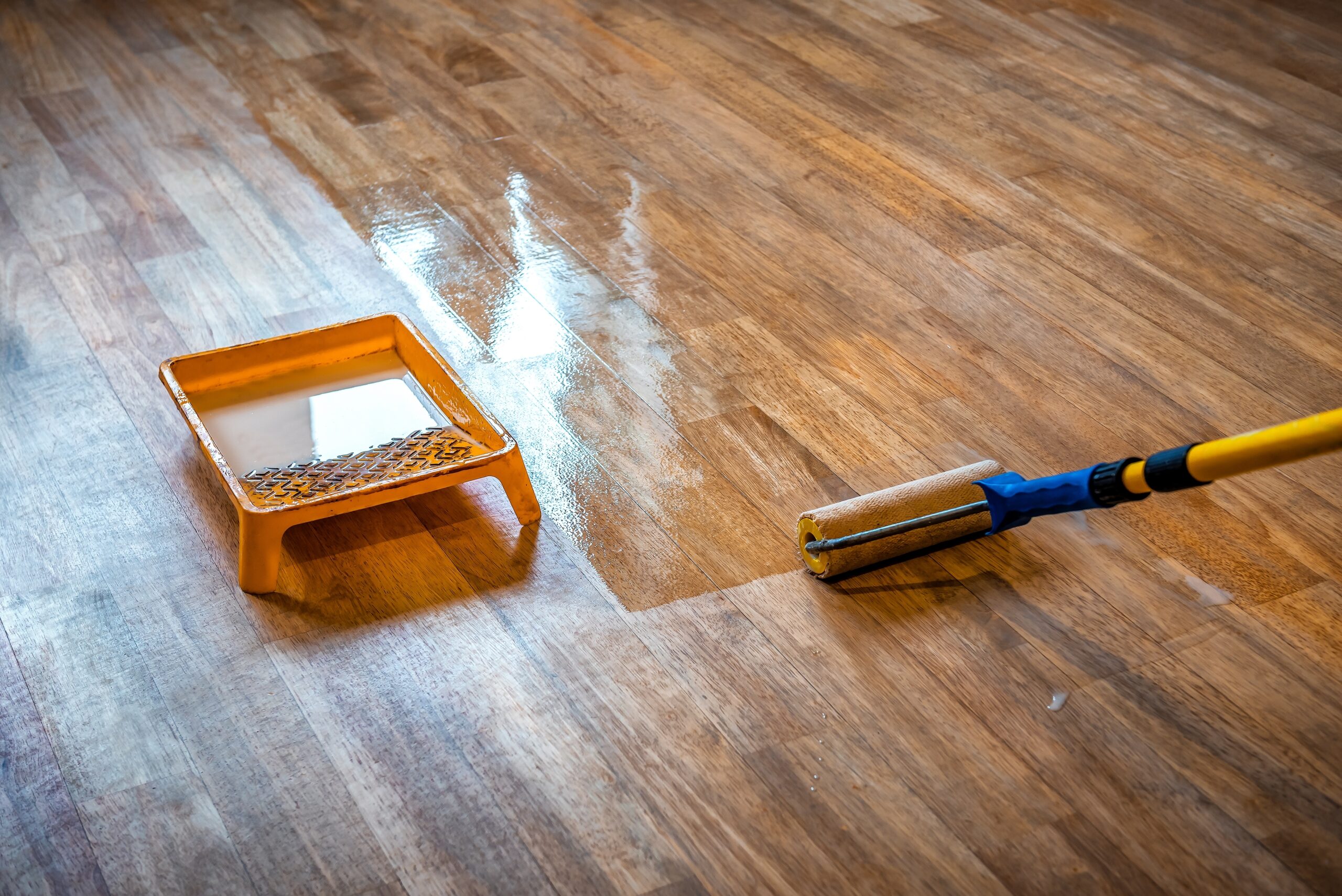 An angled shot showing a roller applicator and an orange paint tray with finish, applying a coat of clear finish to a medium-toned hardwood floor, creating a wet, shiny surface. Hardwood Floor Refinishing Marietta GA by Peachy Flooring Solutions.