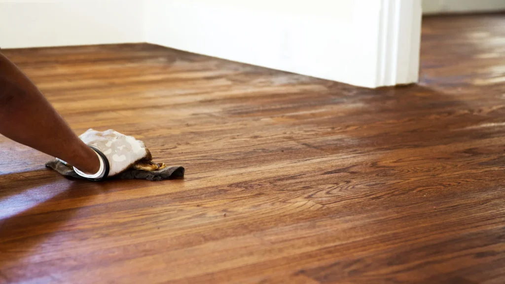 A close-up view of a person wearing a glove applying stain or finish with a rag to a newly sanded hardwood floor, part of the Hardwood Floor Refinishing Roswell GA services by Peachy Flooring Solutions.