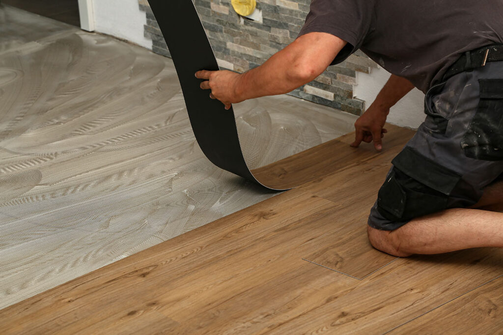 A close-up shot of an installer laying down a plank of light brown, wood-look Luxury Vinyl Plank (LVP) flooring, pulling back the peel-and-stick backing. The newly installed floor is shown next to an area of the subfloor prepared with adhesive or self-leveler. This image showcases a step in the LVP Installation Roswell GA service provided by Peachy Flooring Solutions.