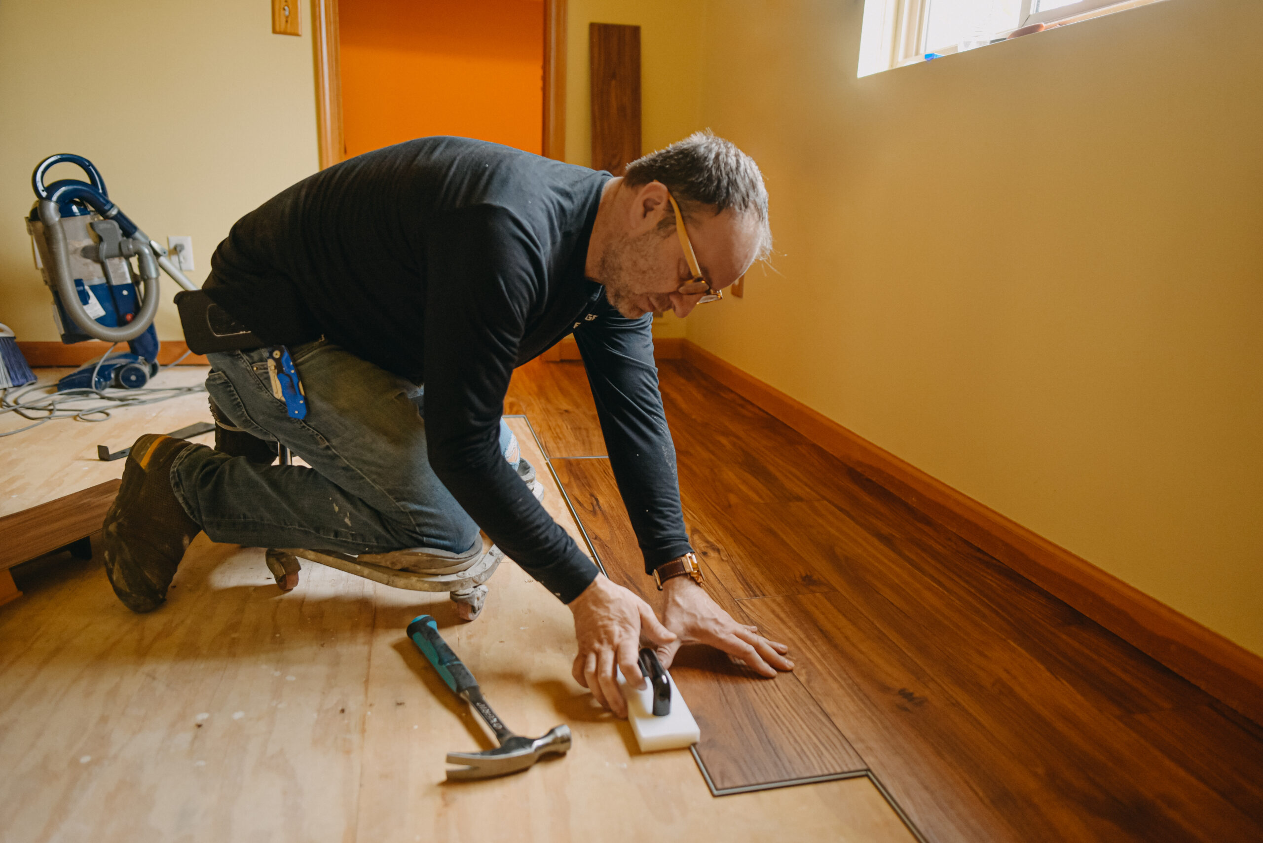 A professional installer kneels on the subfloor, carefully tapping a new plank of wood-look Luxury Vinyl Plank (LVP) flooring into place using a small mallet and tapping block. The room has yellow walls and an orange accent area in the background. The photo depicts the precise LVP Installation Roswell GA process carried out by Peachy Flooring Solutions.