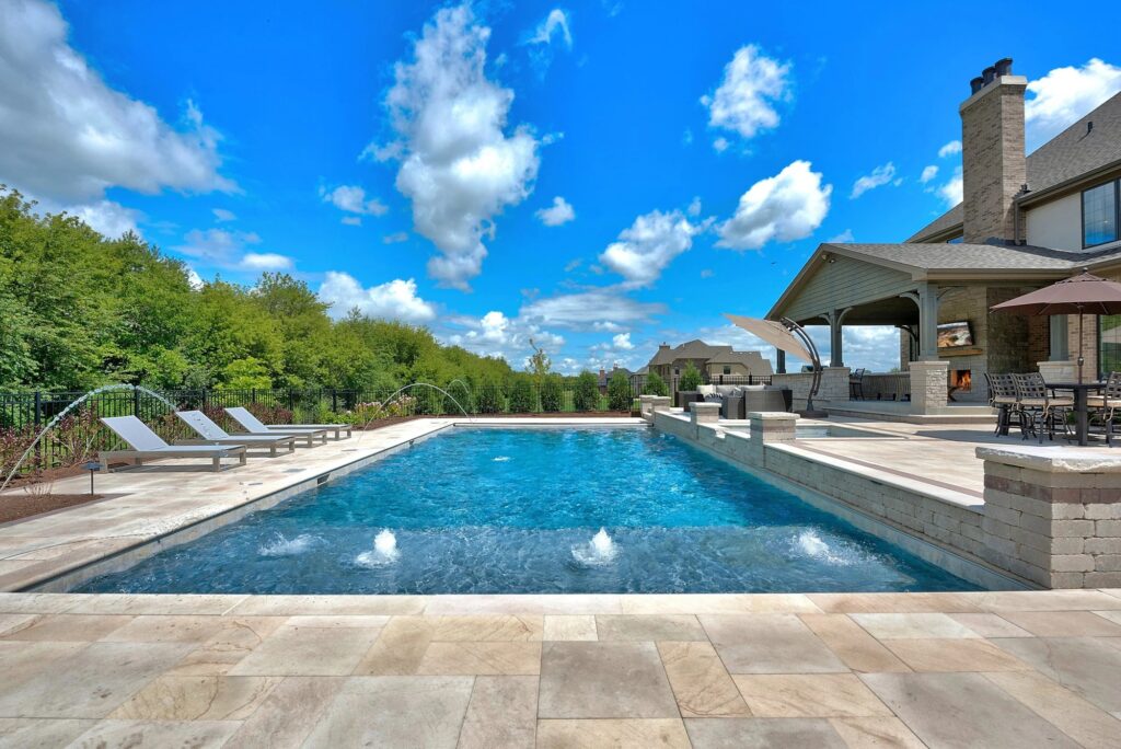 A large, luxurious rectangular custom-built swimming pool with integrated water features and a spacious stone patio, set against a blue sky. This exemplifies Mooresville Hire Custom Pool Construction by Craft Master Pools and Spas.