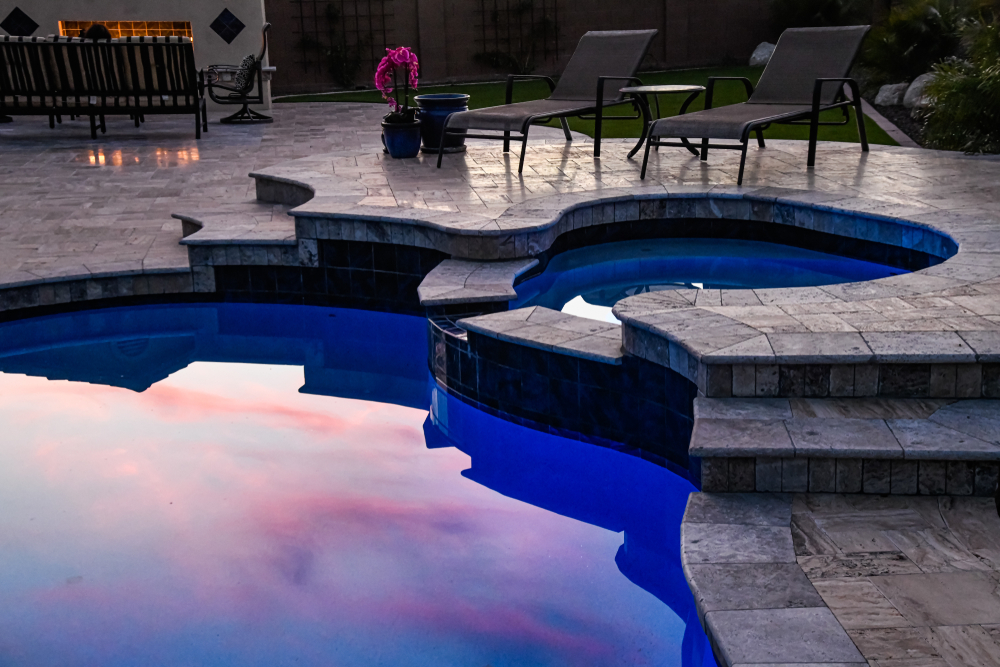 A custom free-form swimming pool with underwater lighting at dusk, showing stone coping and steps. This is an example of Mooresville Hire Custom Pool Construction by Craft Master Pools and Spas.