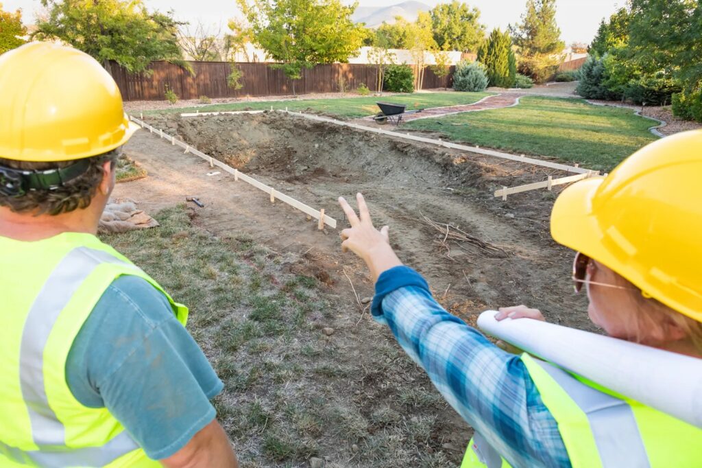 Two pool construction workers in yellow hard hats and safety vests stand over a freshly excavated hole and wood framing for a new pool installation, representing the start of a project by Mooresville Pool Construction Companies by Craft Master Pools and Spas.