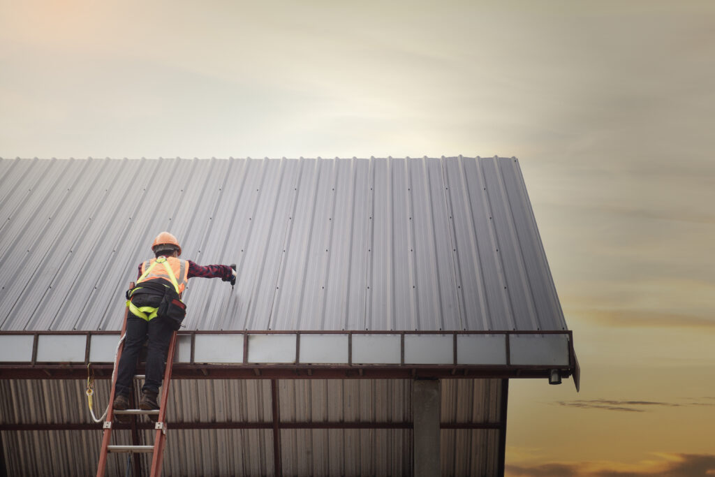 Low-angle shot of a Nashville TN Commercial Roof by Wegner Roofing & Solar, showing a worker in an orange vest and hard hat on a ladder, installing or repairing the corrugated metal roof against a soft, bright sky.