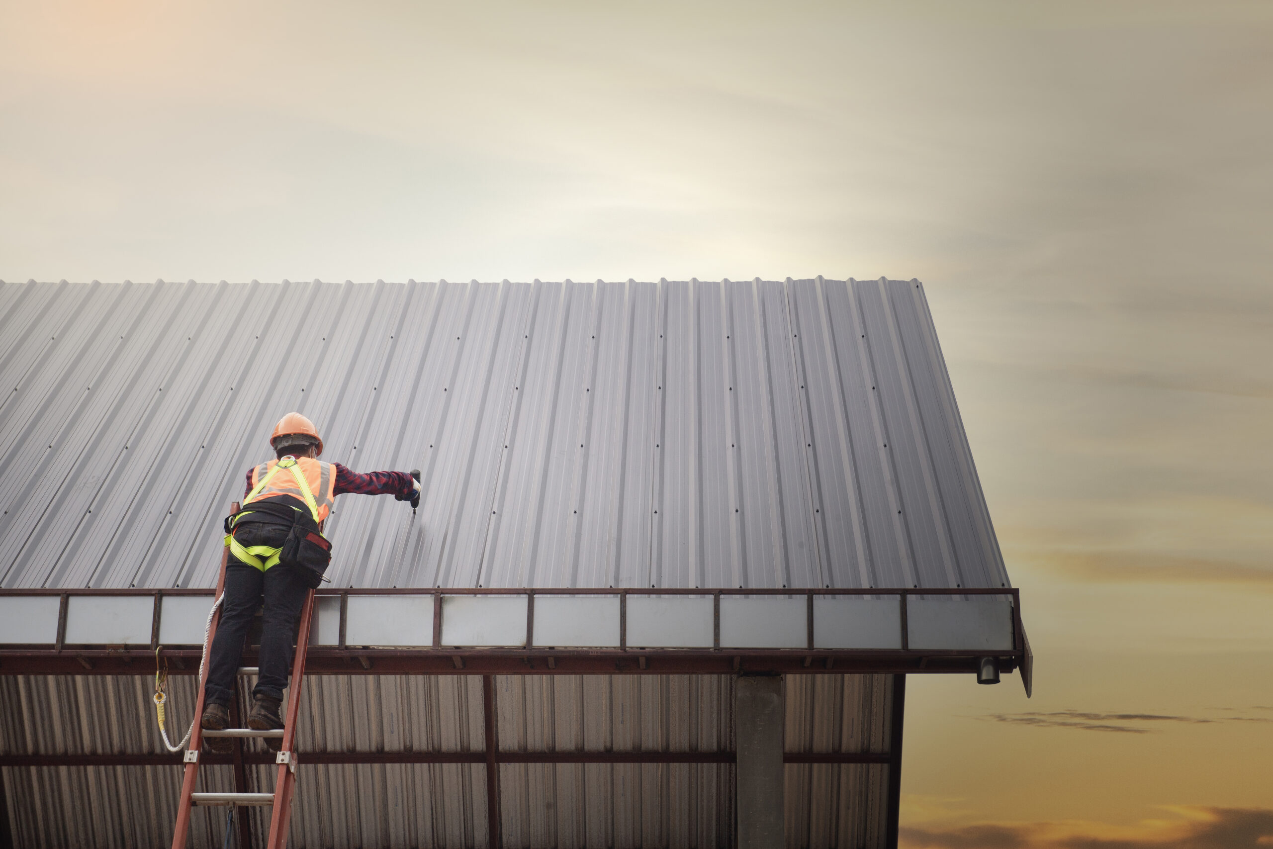 Low-angle shot of a Nashville TN Commercial Roof by Wegner Roofing & Solar, showing a worker in an orange vest and hard hat on a ladder, installing or repairing the corrugated metal roof against a soft, bright sky.