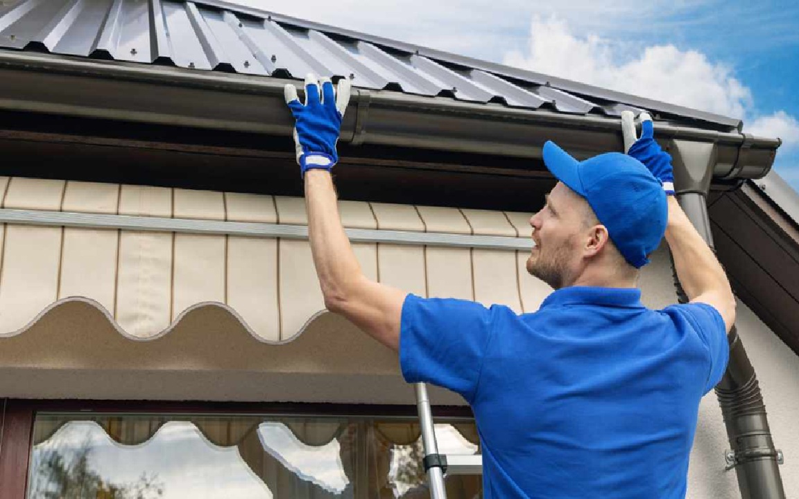 A professional installer from Wegner Roofing & Solar wearing a blue shirt and baseball cap is standing on a ladder, adjusting a newly installed dark brown seamless gutter under a metal roof. This image represents quality Nashville TN Gutter Installation service, showing attention to detail.