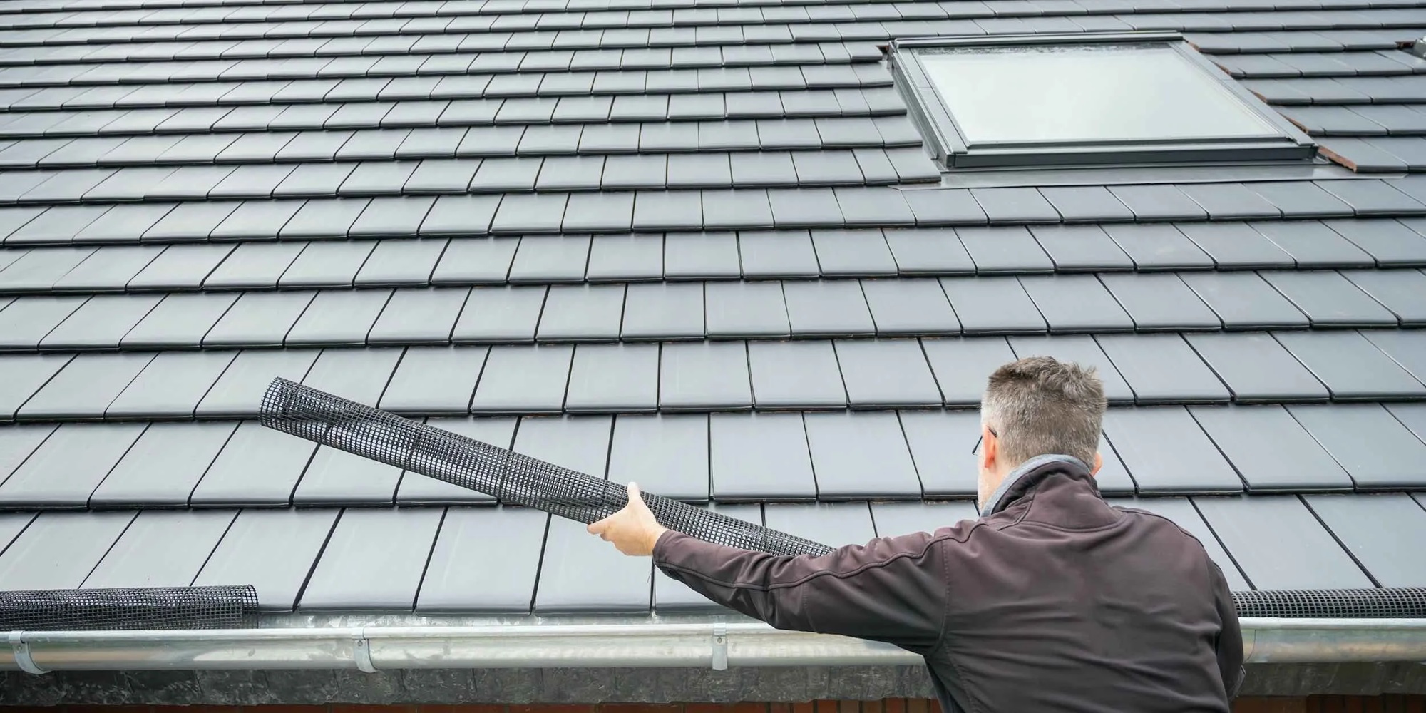 A worker from Wegner Roofing & Solar is preparing to install a black mesh gutter guard system onto a light-colored gutter on a residential roof with gray slate-style tiles and a skylight. This illustrates a step in comprehensive Nashville TN Gutter Installation, focusing on adding protection like gutter guards.