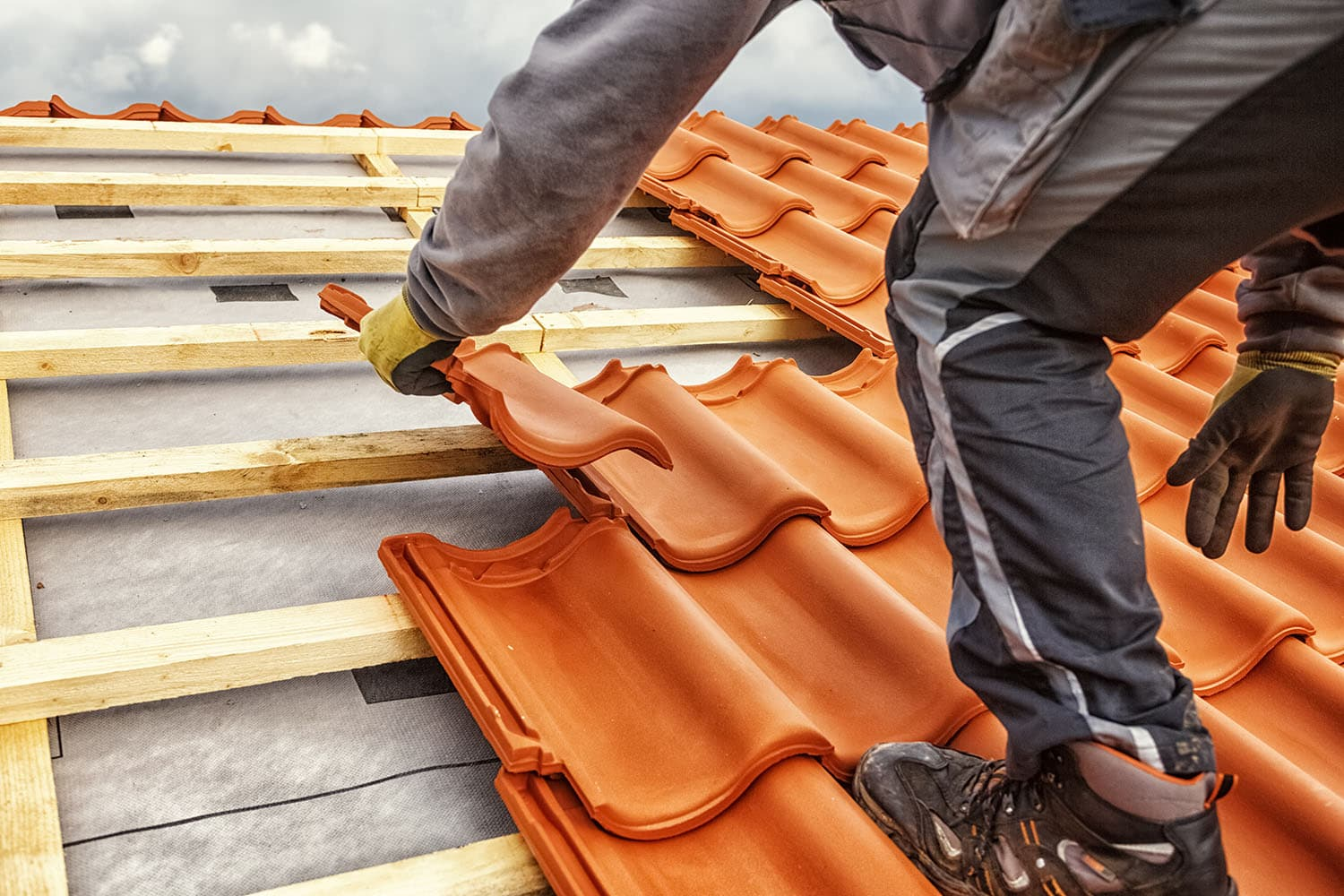 Nashville TN Roofing Replacement by Burish Builder TN: Close-up of a roofer wearing gloves installing orange terracotta tile on a roof over a layer of underlayment and wooden battens.