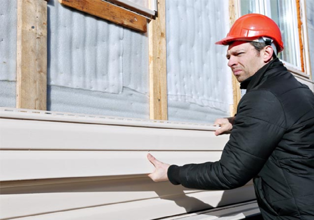 A professional Nashville TN Vinyl Siding Installer by Wegner Roofing & Solar, wearing an orange hardhat and black jacket, is carefully aligning and installing a light beige panel of new vinyl siding on a building's exterior.