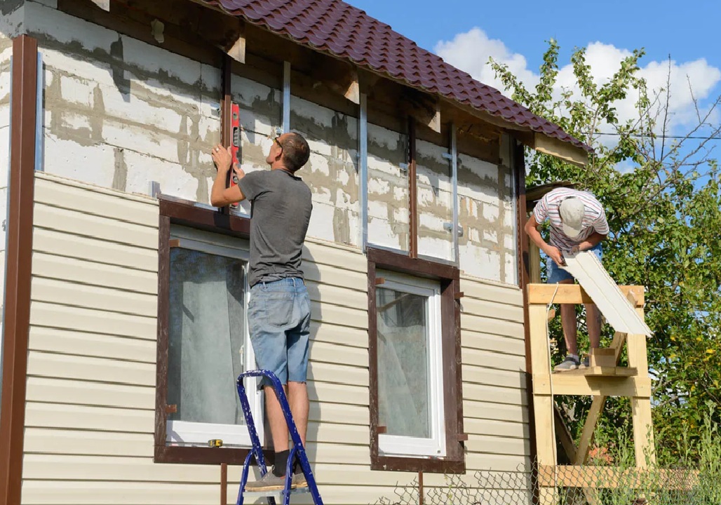 Two Nashville TN Vinyl Siding Installers by Wegner Roofing & Solar working on a residential house. One installer is on a ladder using a level on the metal framing for the new siding, while the other is standing on scaffolding installing a panel of light-colored vinyl siding near a window.