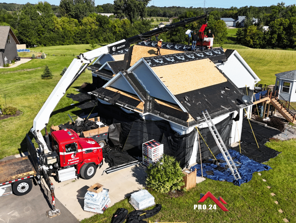 Ramsey MN Storm Damage Repair by Pro24 Contracting, Inc.: A red boom truck lifting materials onto a residential roof being replaced, showing the roof deck with underlayment and stacks of new shingles on the ground.