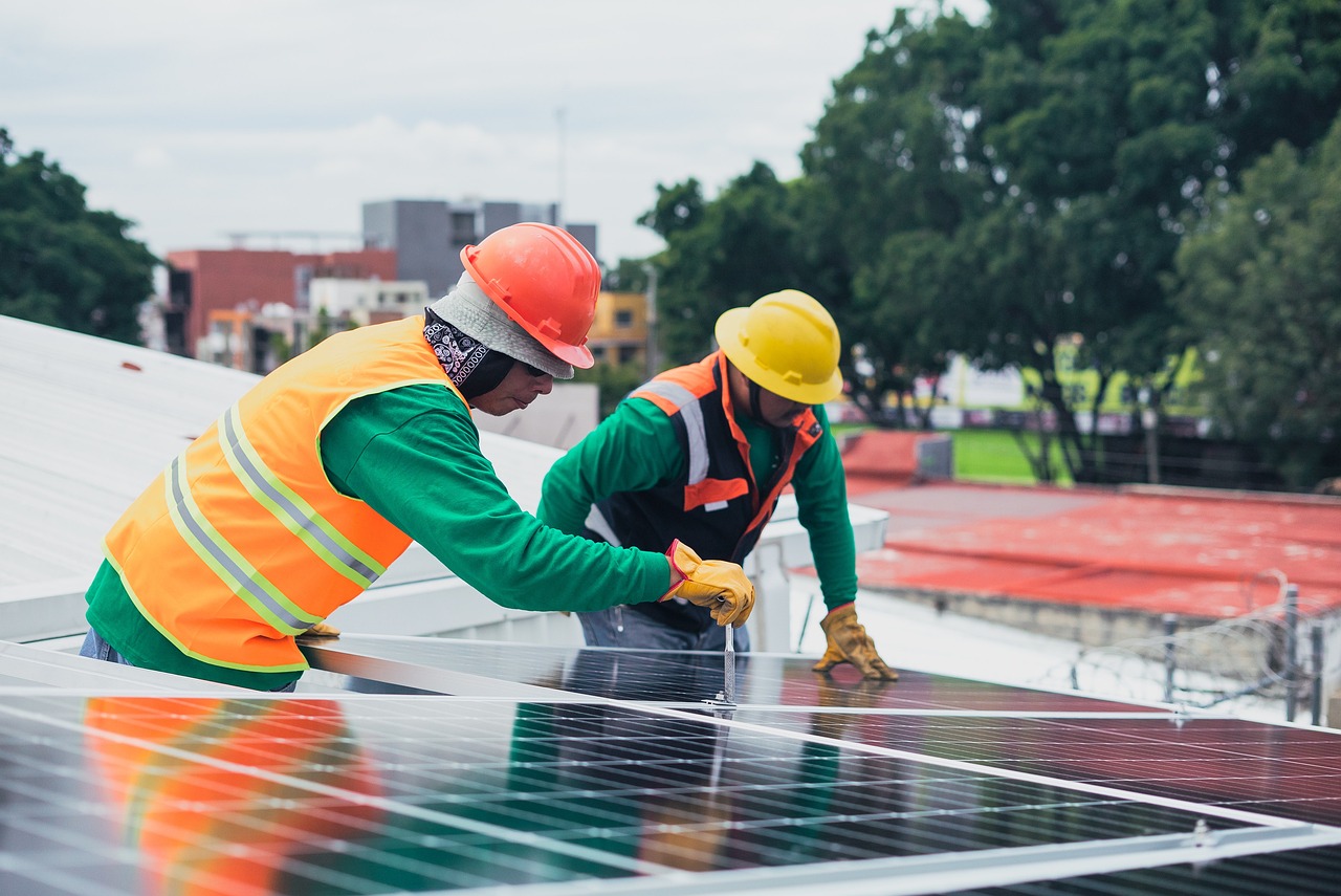 Two installers from Rapid City Solar Companies by Wegner Roofing & Solar working on a rooftop solar panel installation, wearing orange and green safety gear and hard hats.