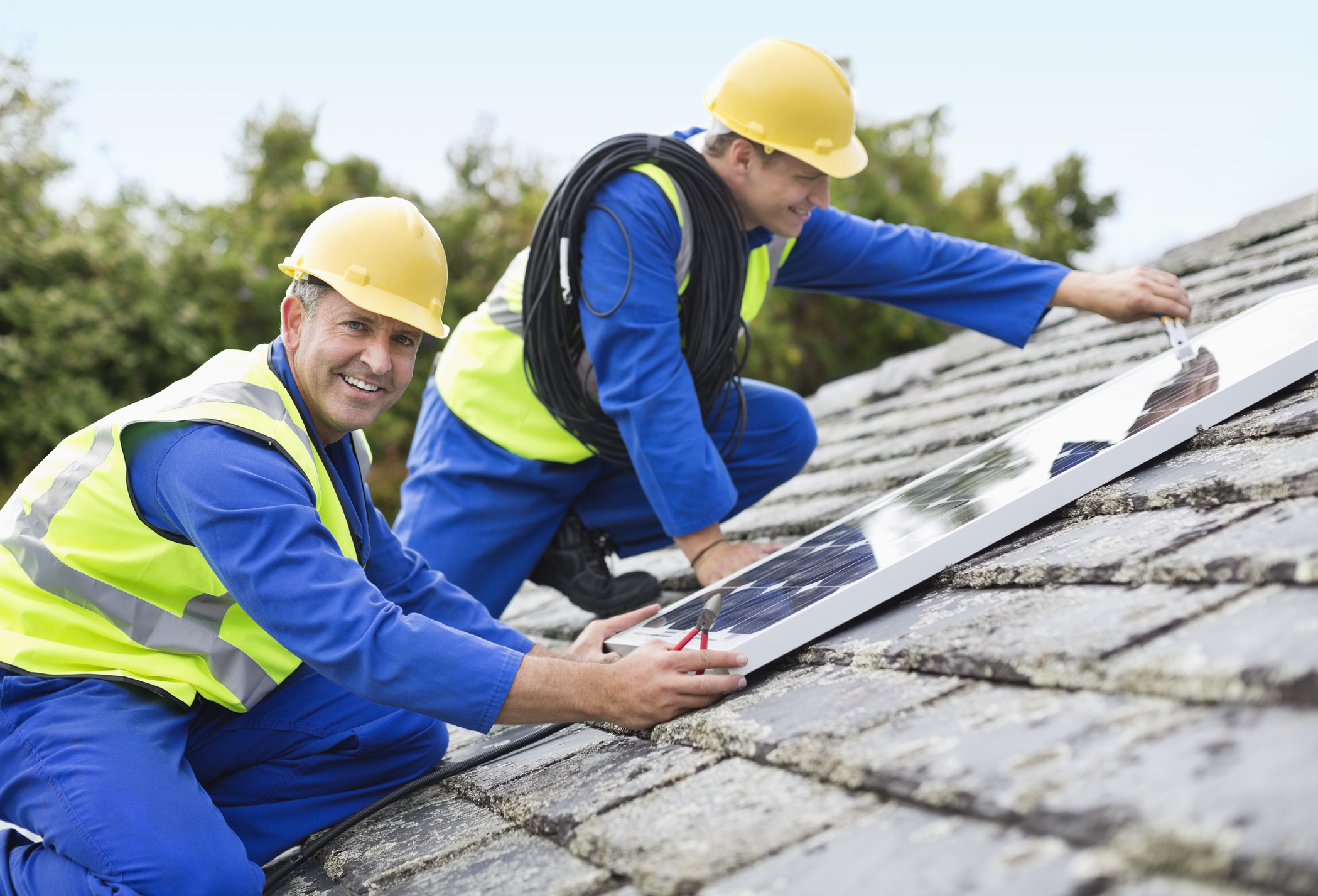 A friendly installer from Rapid City Solar Companies by Wegner Roofing & Solar smiling while his colleague assists in installing a small solar panel on a shingled roof.