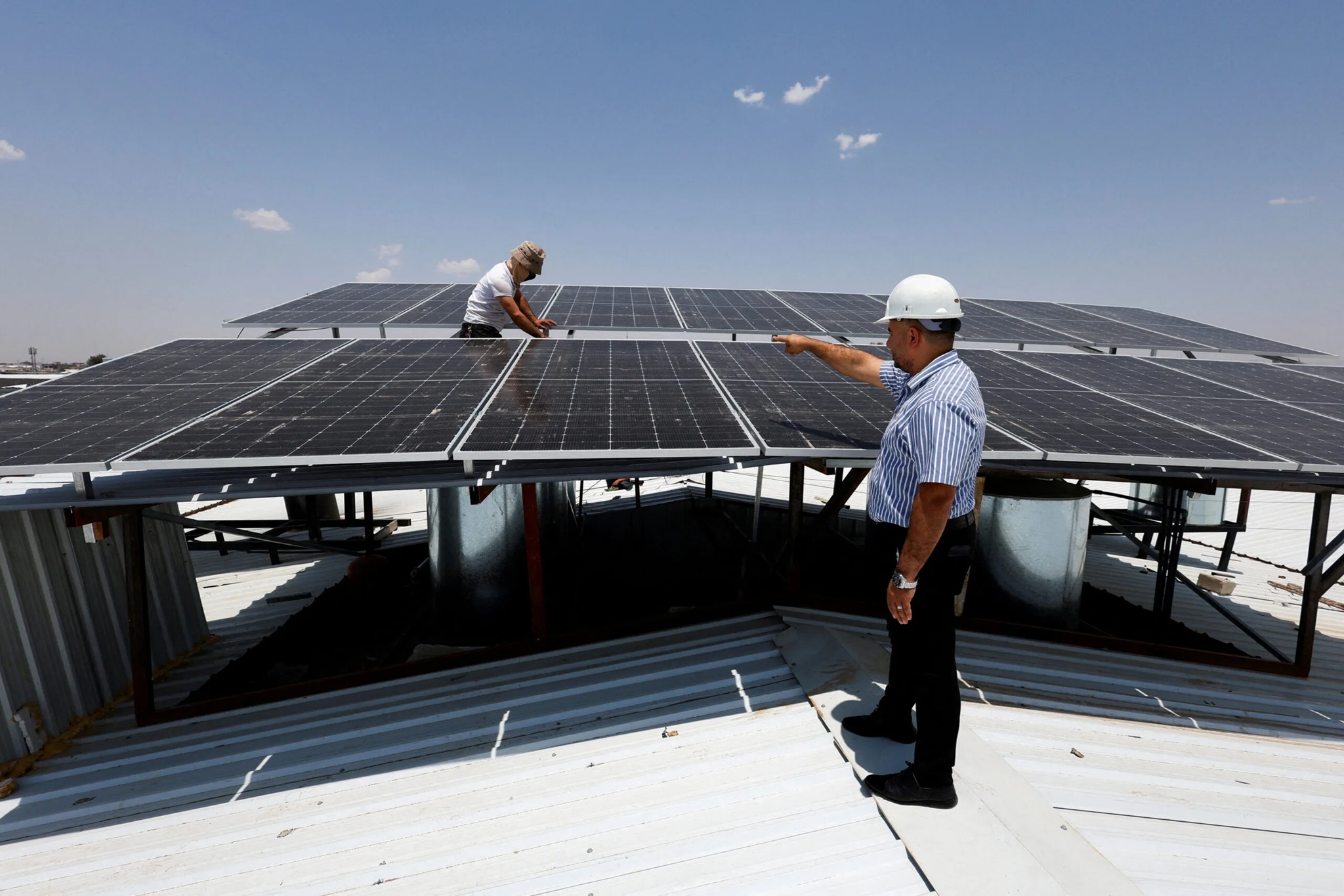 Two workers from the Rapid City Solar Company by Wegner Roofing & Solar are installing or inspecting solar panels on a commercial rooftop. One man in a white hard hat and striped shirt is pointing towards the elevated solar array while another worker in a white t-shirt and hat works directly on the panels above. The panels are elevated on metal framing above a white metal roof under a bright, sunny sky.