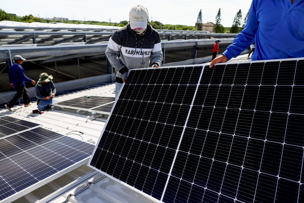 Close-up of a worker, part of the team at Rapid City Solar Company by Wegner Roofing & Solar, handling a large, black solar panel on a rooftop. The worker is wearing a grey and white hoodie and a cap. Another worker in a blue shirt is partially visible to the right, assisting with the panel. Other workers and installed solar panels are visible in the background on the white corrugated roof.
