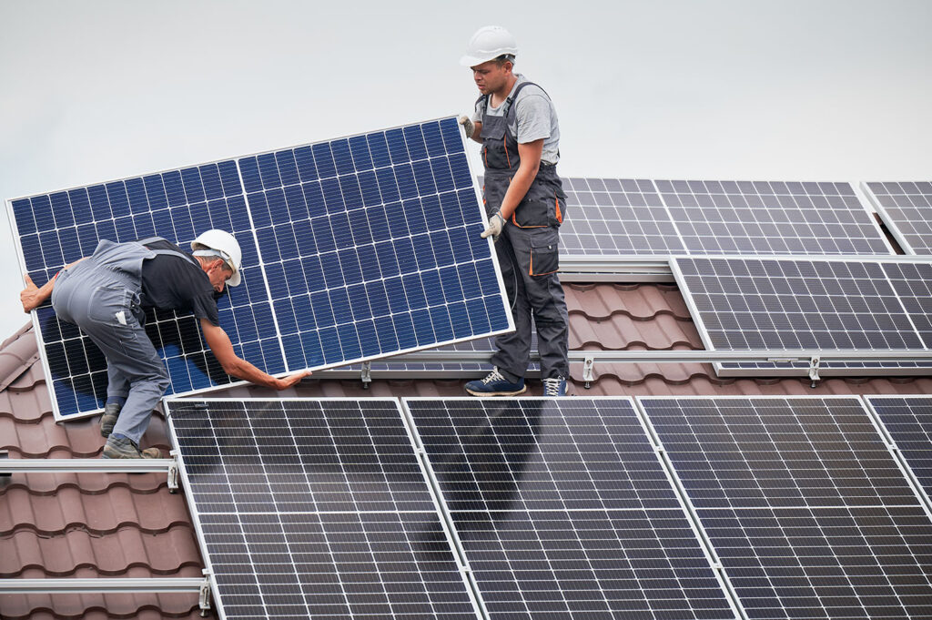 Two installers in work overalls and white hard hats performing a Rapid City Solar Installation by Wegner Roofing & Solar. They are carefully maneuvering a large blue solar panel into place on a brown tiled roof that is already partially covered with other installed solar panels.
