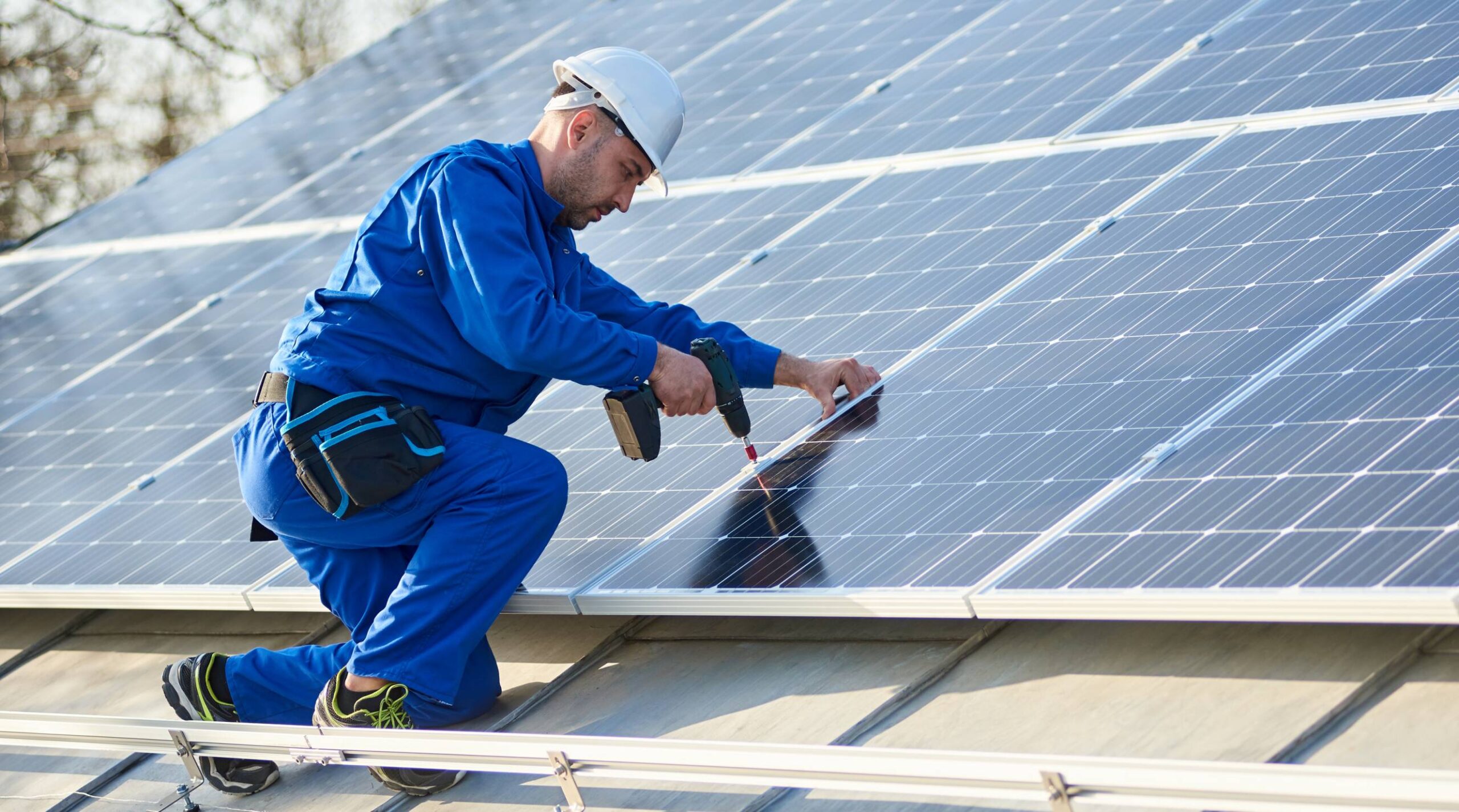 A professional solar installer in a blue jumpsuit and white hard hat uses a drill to secure a solar panel on a rooftop. This image represents the Rapid City Solar Installation Near Me service offered by Wegner Roofing & Solar.