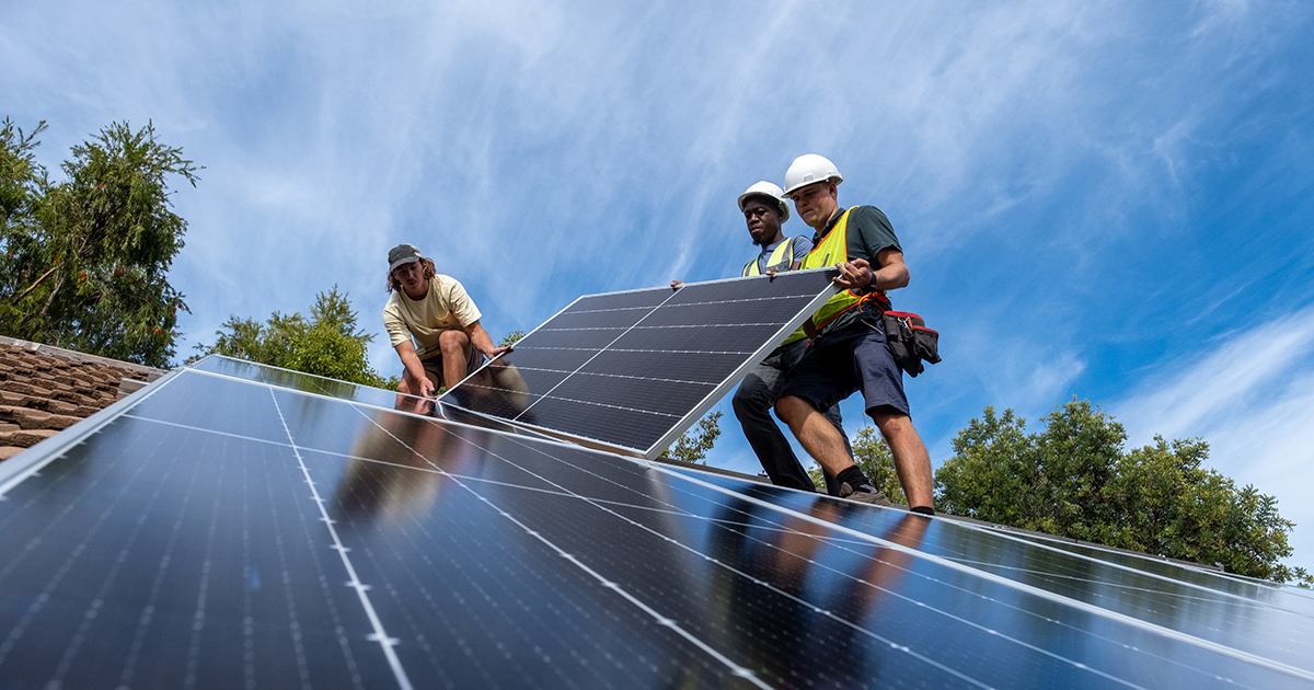 Three technicians performing a Rapid City Solar Installation by Wegner Roofing & Solar. Two workers, one wearing a hard hat and safety harness, are lifting and positioning a large black solar panel onto a terracotta-tiled roof next to already installed panels, set against a bright blue sky with scattered clouds.