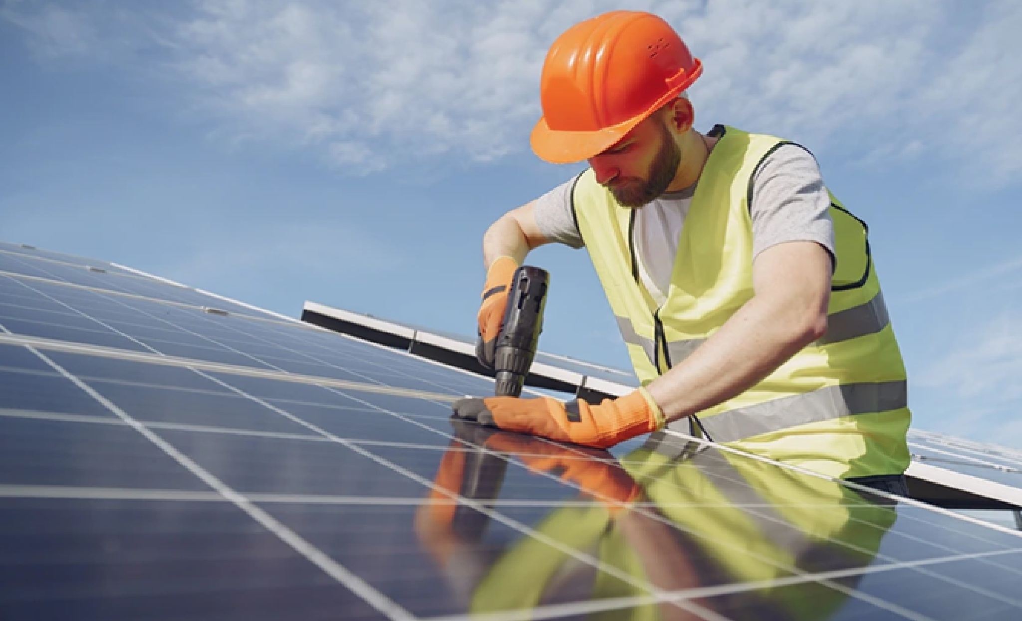 Close-up of a Rapid City Solar Installer from Wegner Roofing & Solar wearing an orange hard hat and safety vest, using a power drill to secure a solar panel on a rooftop array under a bright blue sky.