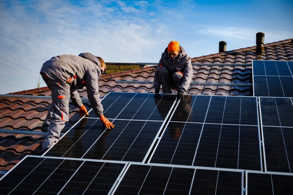 Rapid City Solar Panel Installers from Wegner Roofing & Solar are working on installing solar panels on a brown tiled roof under a bright blue sky. Two installers in work clothing are shown kneeling and positioning the dark photovoltaic panels.