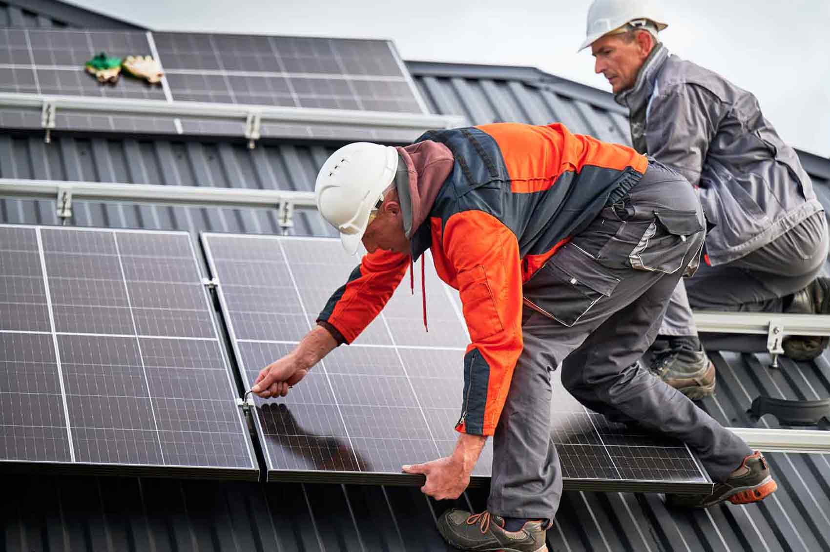 Two Rapid City Solar Panel Installers from Wegner Roofing & Solar, wearing hard hats and safety gear, are securing a dark solar panel onto a black metal roof. One installer is bent over, making an adjustment, while the other is kneeling behind him.