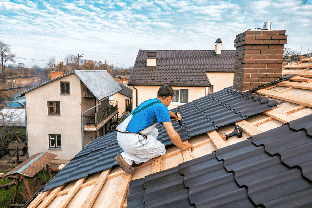 A roofing contractor installing dark, interlocking metal roof panels onto a residential home's exposed wooden frame under a bright, cloudy sky. Image for Residential Roofing Services Forest Acres SC by Burish Builders Columbia.