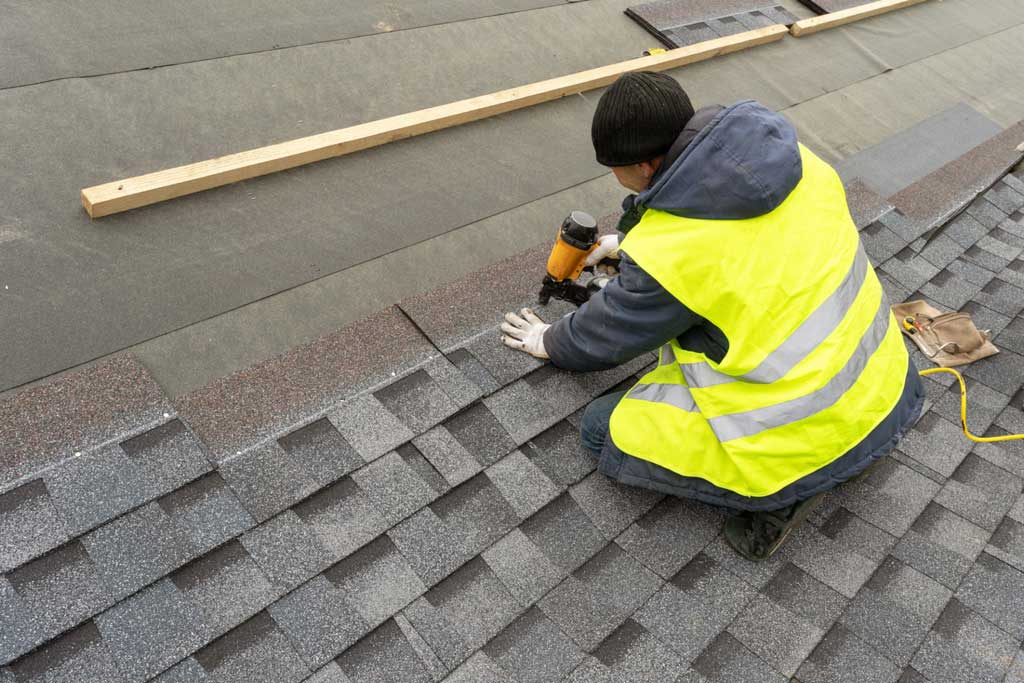 Expert Residential Roofing Services Lexington SC by Burish Builders Columbia: A worker installs gray asphalt shingles onto a roof deck, using a nail gun and wearing a high-visibility vest and dark winter cap.