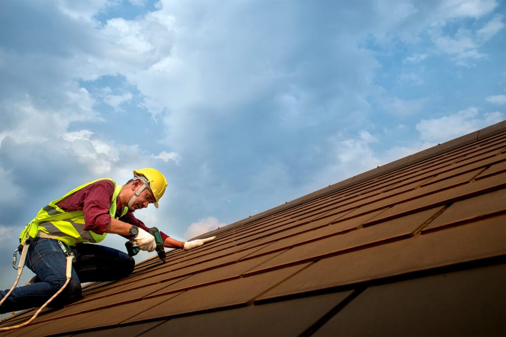 A roofer in a yellow safety vest and hard hat installing brown metal roofing tiles, representing high-quality Residential Roofing Services Sunset Beach NC by Burish Builders Wilmington.