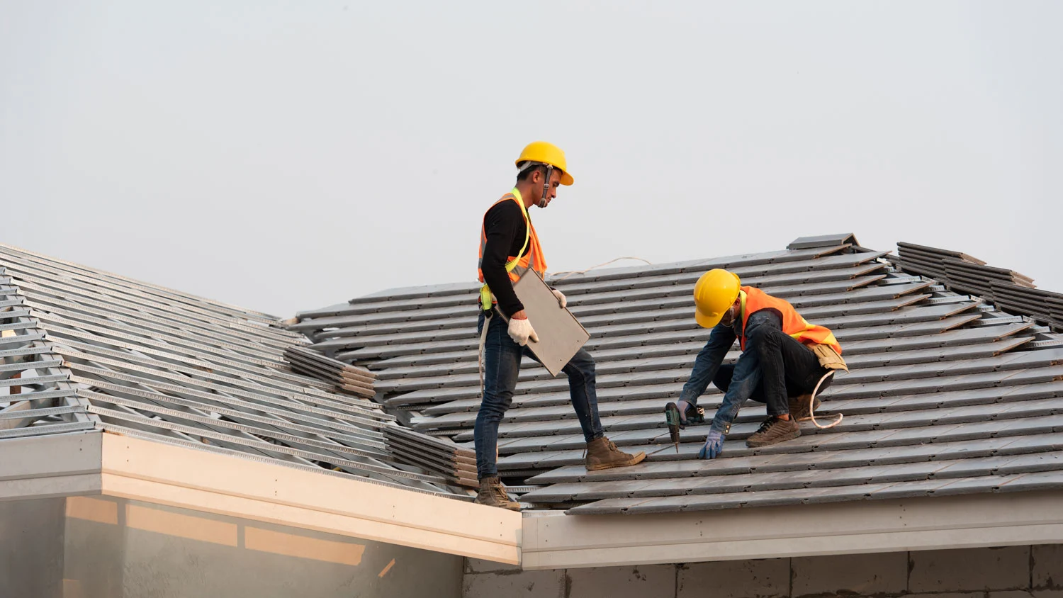 Two construction workers, wearing safety vests and hard hats, are installing gray roof tiles on a residential structure with an exposed metal frame. One worker stands, holding a tile, while the other kneels and uses a drill. This work is typical of a Roofing Company Forest Acres SC project by Burish Builders Columbia.