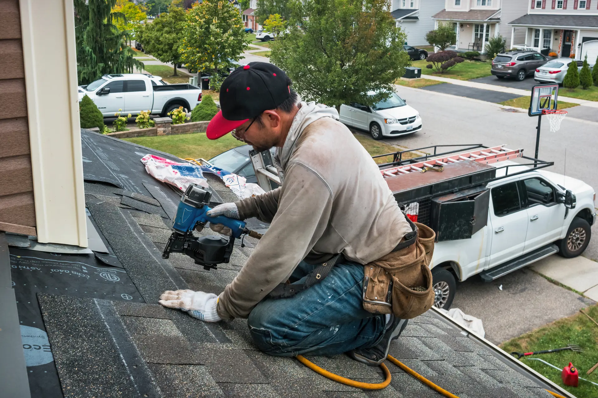 A roofing professional, wearing a red and black cap and a tool belt, kneels on a partially installed roof and uses a pneumatic nail gun to fasten shingles. The surrounding residential neighborhood and parked vehicles are visible. Roofing Installation Lexington SC by Burish Builders Columbia.