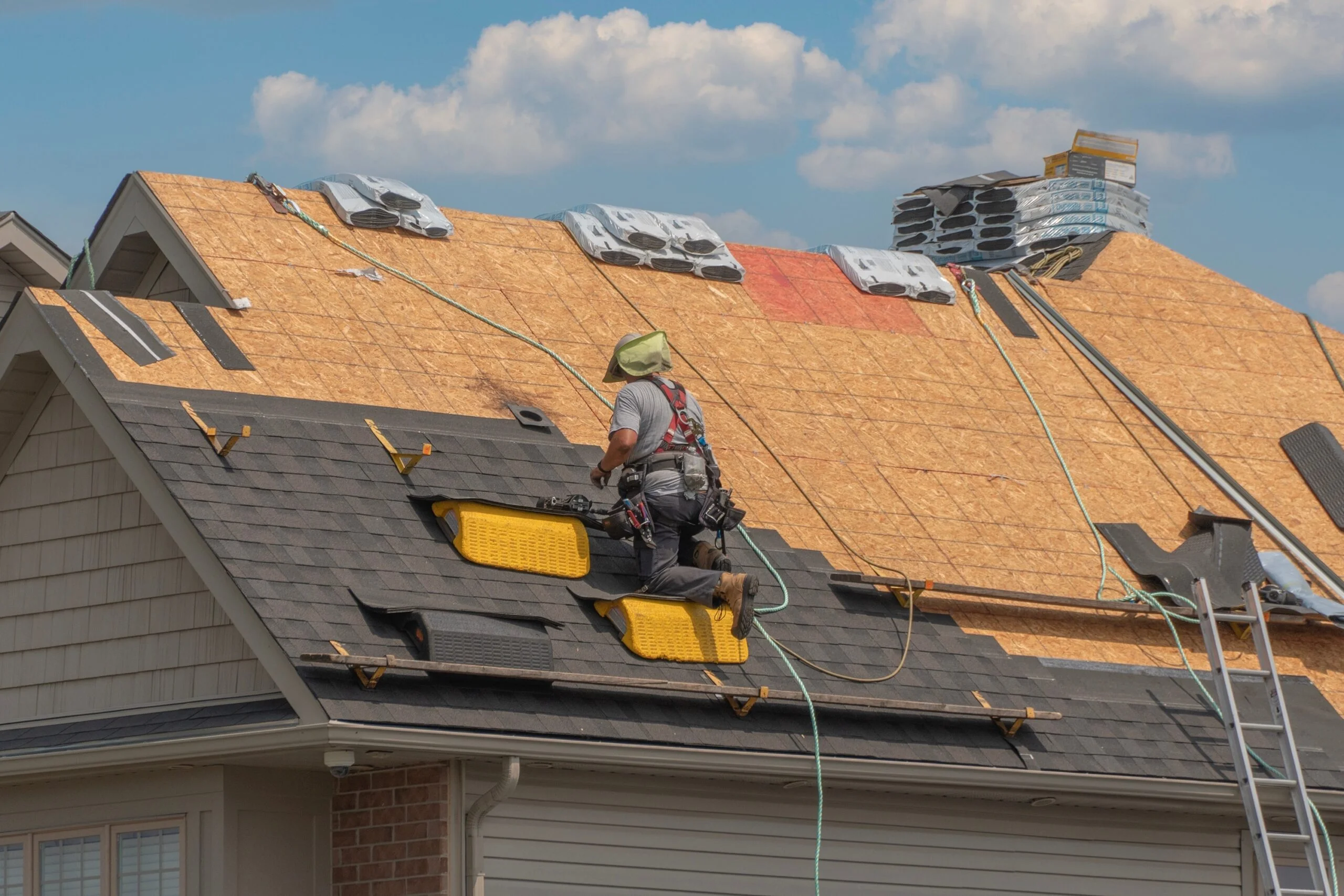 A wide-angle shot of a residential roof undergoing a Roofing Replacement Lexington SC by Burish Builders Columbia. The roof decking is exposed on the upper section, while the lower section shows new, dark shingles being installed, with stacks of material and a worker secured by a safety harness.