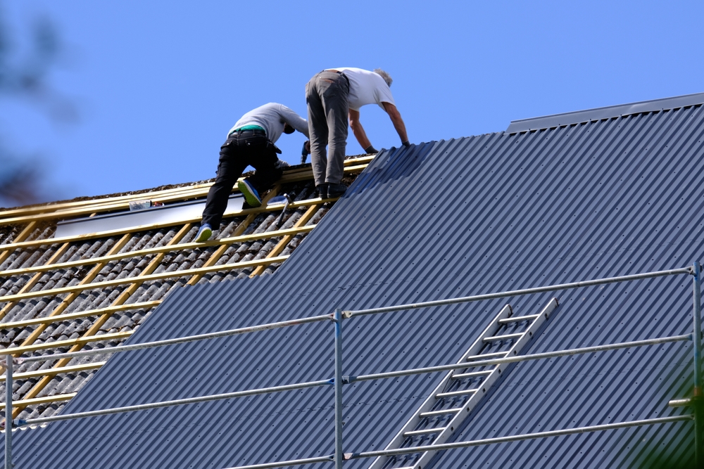 Two roof workers installing blue corrugated metal roofing panels on a steep residential roof, with some of the wooden understructure exposed. Scaffolding with a ladder is visible in the foreground. This image is of a Roofing Replacement Sunset Beach NC being performed by Burish Builders Wilmington.