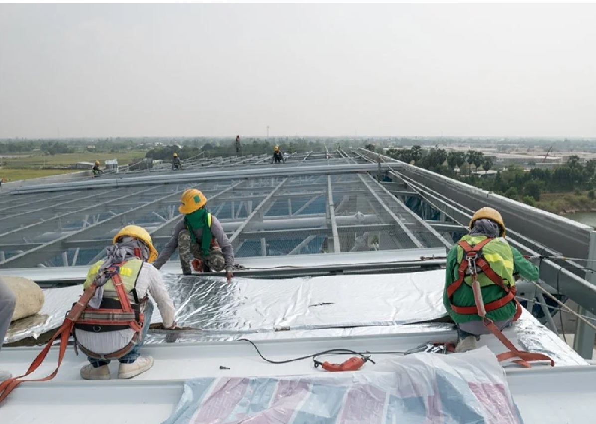 Four Spearfish Commercial Roofers by Wegner Roofing & Solar are installing a reflective roofing membrane or insulation on a large metal-framed commercial building, wearing hard hats and fall protection harnesses.