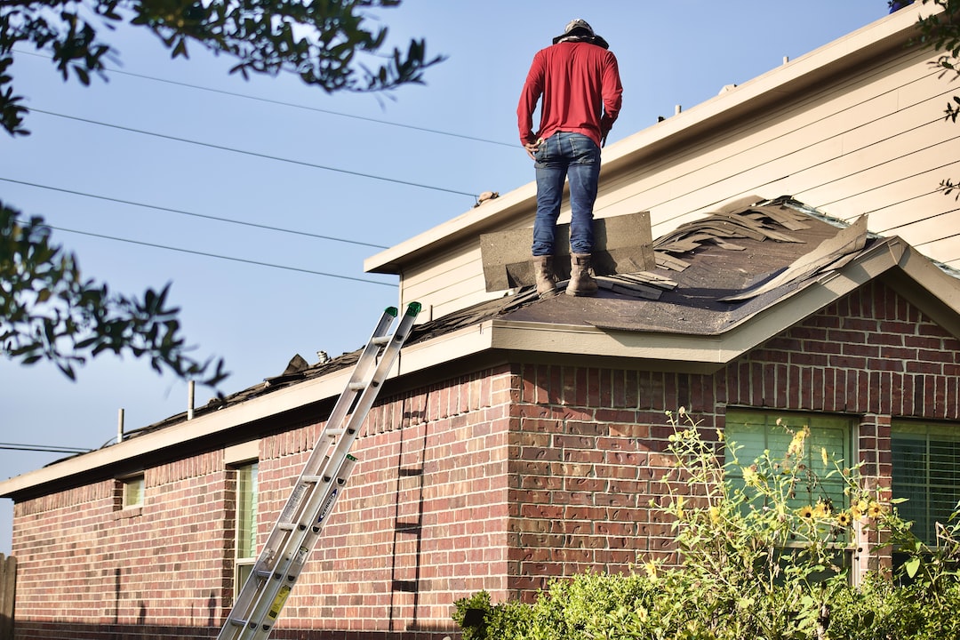 A roofer from Spearfish Emergency Roofing Services by Wegner Roofing & Solar stands on a damaged residential roof, assessing the wind or storm damage to the shingles, with a ladder leaned against the house nearby.
