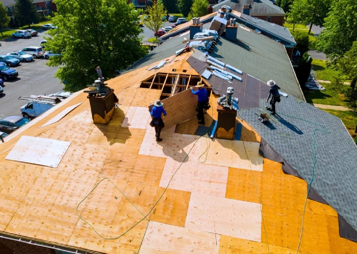 Spearfish Roofing Companies by Wegner Roofing & Solar installing a new roof on an apartment complex. Multiple construction workers in blue shirts are laying plywood sheathing and base layers on the large pitched roof. Part of the roof has new dark shingles already installed, while the rest shows the exposed wood deck and underlying structure, highlighting the reroofing process.