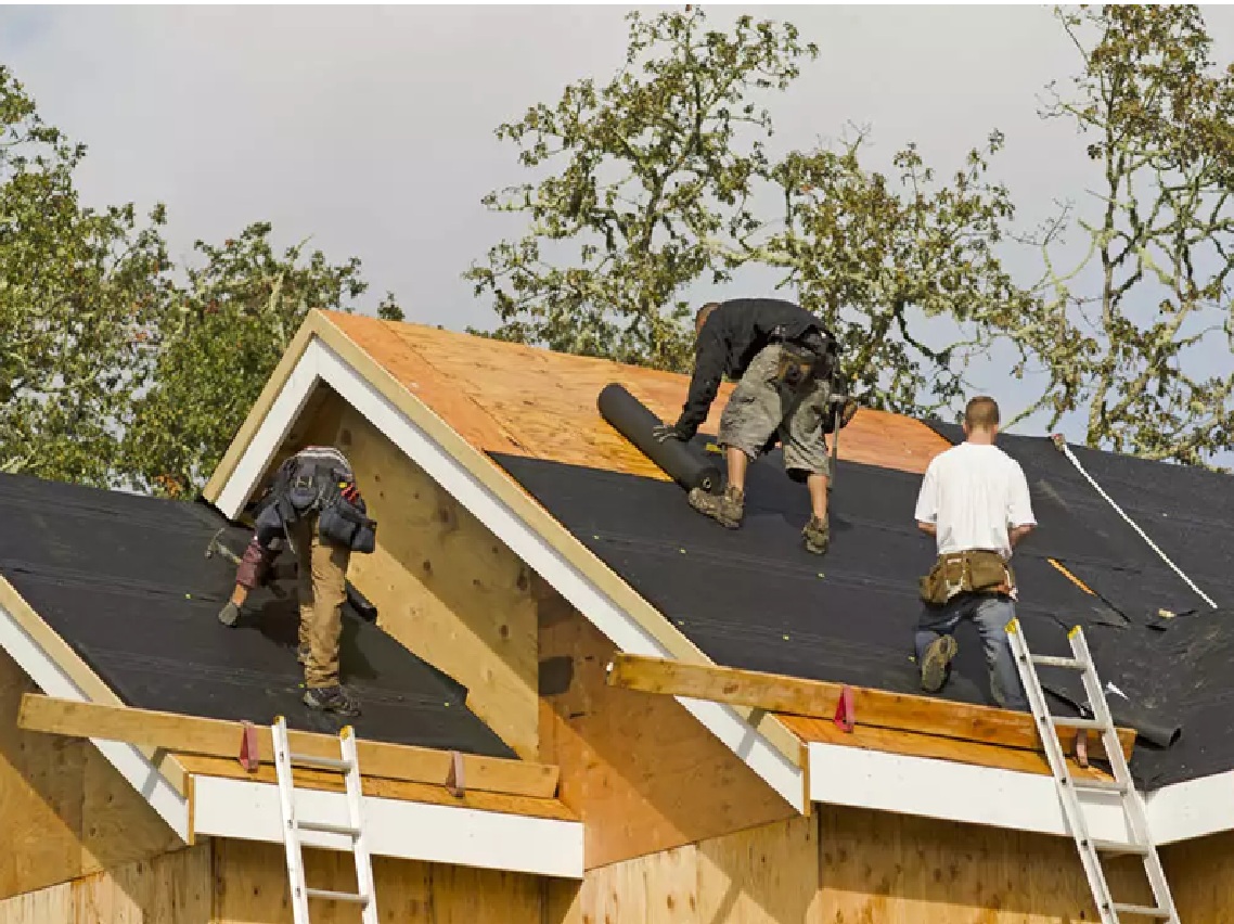 Spearfish Roofing Companies by Wegner Roofing & Solar working on a residential roof replacement. Two roofers are installing a black synthetic underlayment over the exposed wood sheathing on a gable roof section. A third roofer sits nearby, with a ladder propped against the house in the foreground, demonstrating the critical steps of a new roof installation.