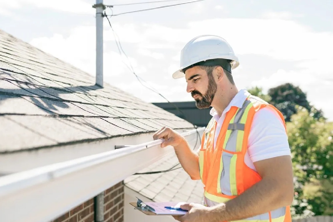 A Spearfish Roofing Contractor from Wegner Roofing & Solar inspecting a white gutter and roofline while wearing a hard hat and safety vest, holding a clipboard.